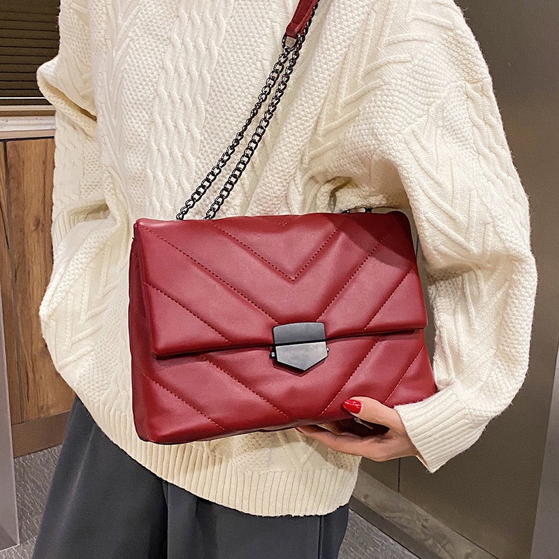 Borsa trapuntata rossa con cuciture a V, tracolla a catena e chiusura nera, portata sulla spalla con una mano sul bordo