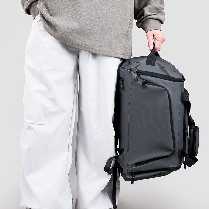 Persona con maglione e pantaloni chiari che tiene lo zaino grigio per la maniglia superiore, vicino al corpo