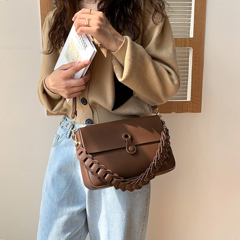 Donna con cardigan beige e jeans chiari, con un libro in mano, porta la borsa marrone allโaltezza del fianco